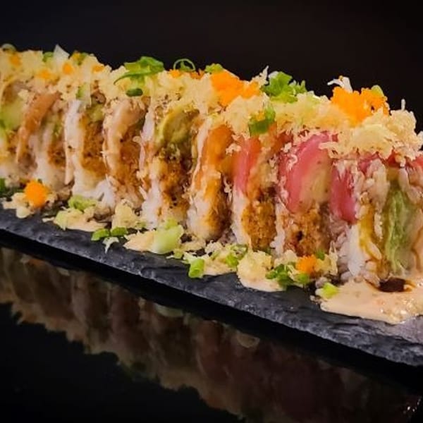 A sushi roll with avocado, pink fish, and orange roe, drizzled with sauce on a dark slate.