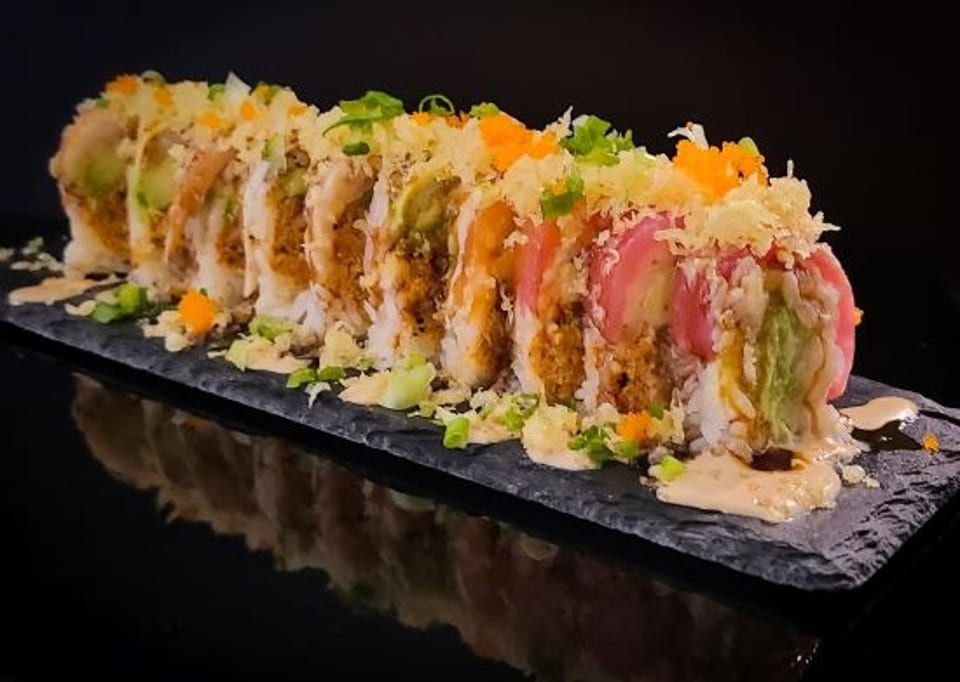 A sushi roll with avocado, pink fish, and orange roe, drizzled with sauce on a dark slate.