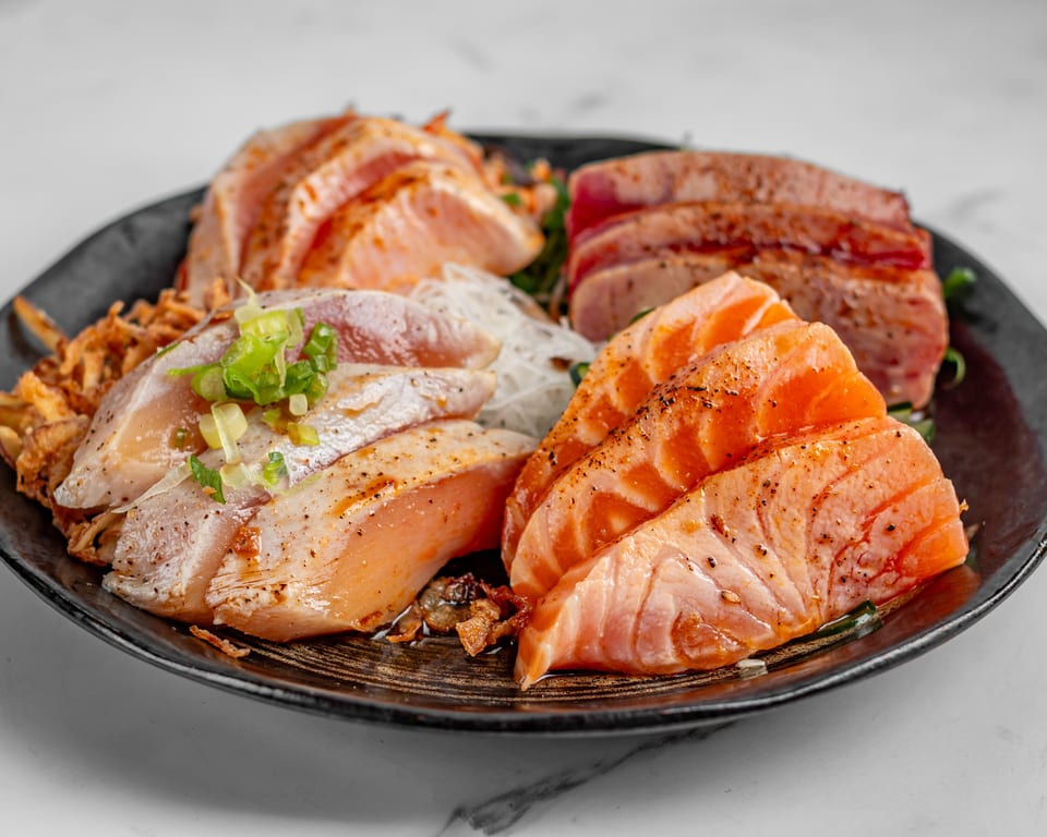 A black plate filled with various cuts of seared salmon and tuna sashimi, garnished with scallions.