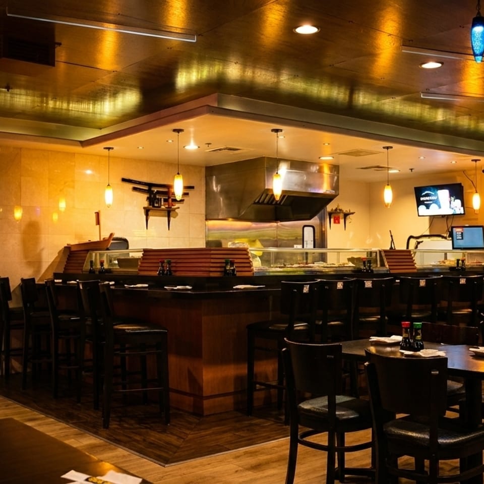 Interior of a sushi restaurant with a long bar, dining tables, and a TV displaying food.