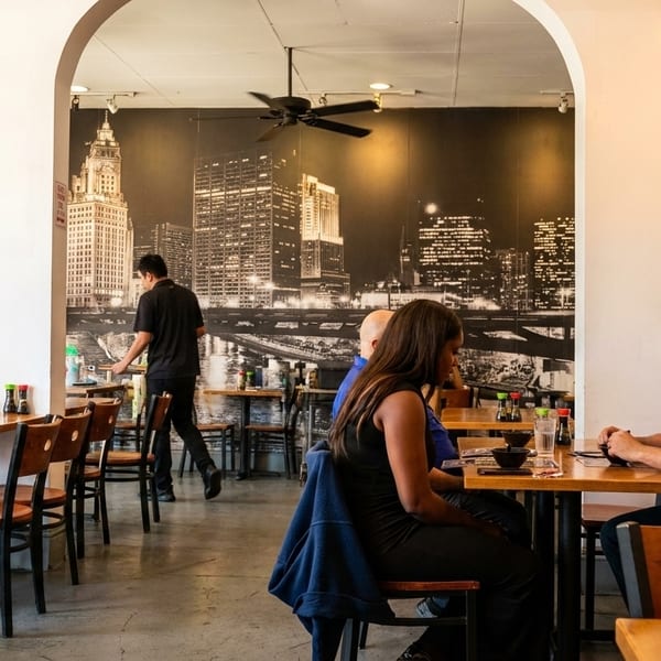 Diners seated at wooden tables in a restaurant with a city skyline mural and a server walking by.