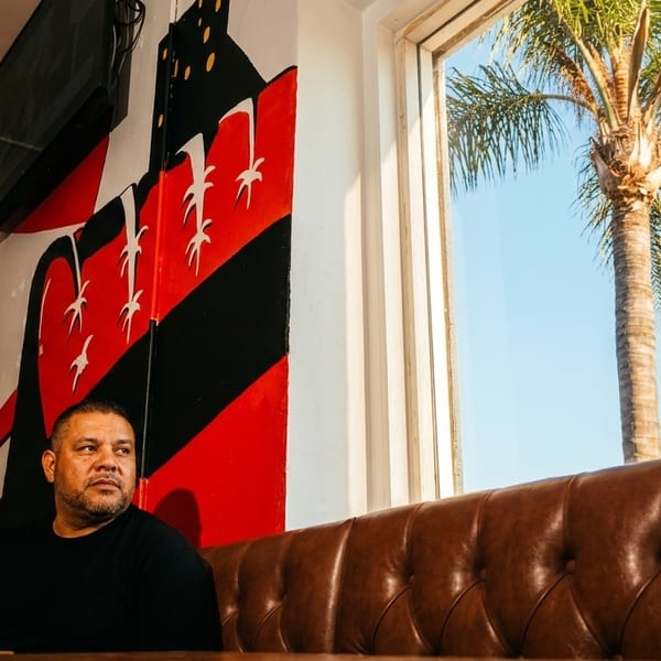 Man sitting on a brown leather booth next to a window with a palm tree outside.