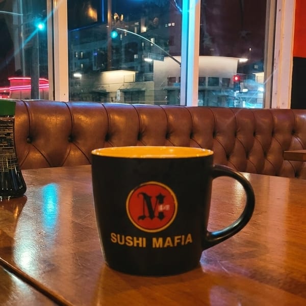 Sushi Mafia mug and soy sauce on a wooden table with a brown leather booth and city lights outside.